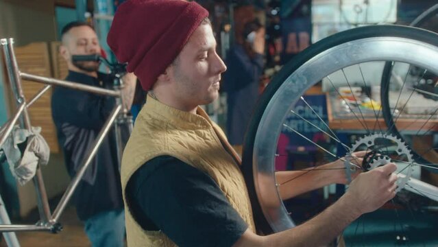 Young male mechanic holding bicycle wheel and examining as it spins, his colleagues working in the background in a repair shop. Side view, medium shot