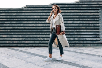 Obraz premium Full length of a walking middle-aged businesswoman with a folder talking on a mobile phone