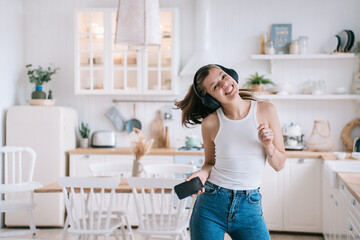 Beautiful American brunette in casual clothes dancing in the kitchen using headphones holding the phone looking at the camera smiling enjoying the weekend at home waiting for her man - obrazy, fototapety, plakaty