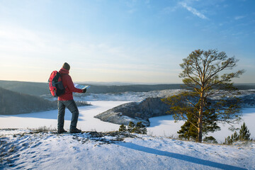 A man standing on top of a mountain with a backpack, using map at winter season.  Winter outdoor hiking.