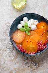 Blue bowl with sliced persimmon, mozzarella balls and pomegranate, vertical shot on a beige granite background, elevated view