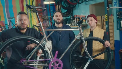 Team of young professional mechanics standing by DIY custom bike and posing on camera together in the workshop. Group portrait, zoom shot