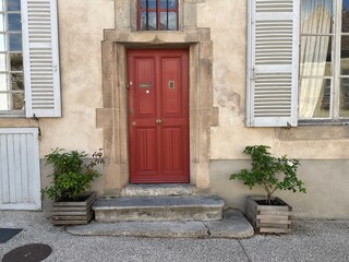 Entrance to a French house with a red door and roses in wooden planters. Windows with wooden...
