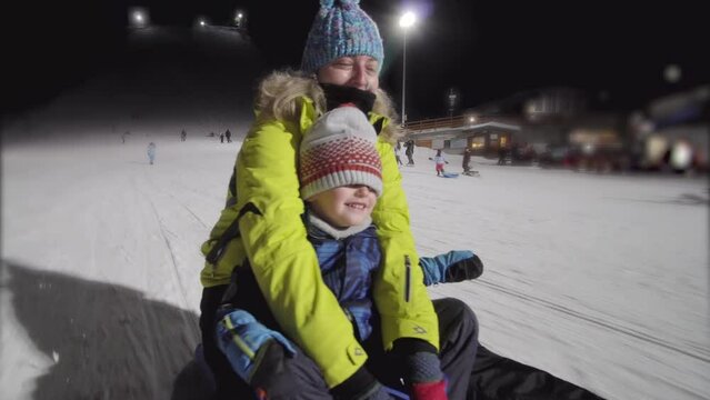 Mother and son on a sledge having fun. Sledging at night on a lit piste.