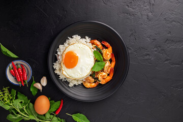 Stir-fried shrimp with basil Thai street food arranged on a black plate with a fried egg Thai spicy food Placed on a dark black table. Top view.