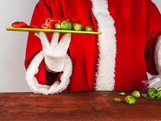 Santa Claus in a traditional costume holds a cutting board with fresh red sweet pepper and broccoli.