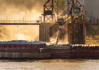 Tugboat pushing freight barges past grain loading dock with dust illuminated by the sun in East St Louis Illinois - obrazy, fototapety, plakaty