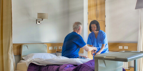 Asian female doctor helping elderly patient in hospital bed. Rehabilitation and retirement concept
