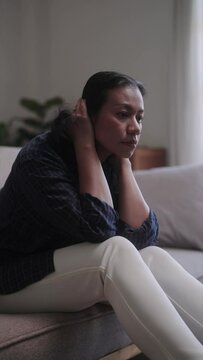 Stressed Asian middle-aged woman at home dealing with health concerns. Overcoming anxiety and seeking relaxation in the living room on her day off.