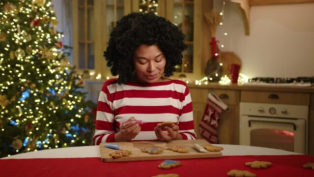 Young black woman painting handmade cookies in pink using tube with food coloring, looking with admiration at bakery. Festively decorated kitchen, garlands. High quality 4k footage