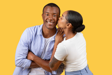 Black woman kissing man on cheek, he smiles at camera - obrazy, fototapety, plakaty