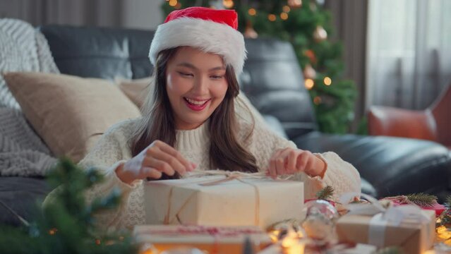 Happy asian woman wearing Santa hat decorates a box wrapped in crab paper with a fir branch, Female hands decorating Christmas present, Christmas gifts wrapping, preparation for holiday season.