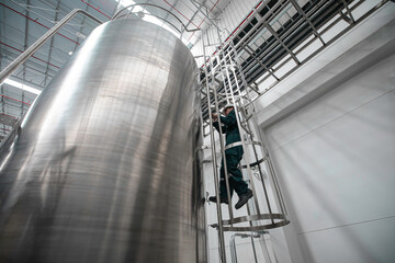 The male worker down the ladder is for inspection stainless steel tank chemical. - obrazy, fototapety, plakaty