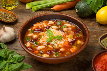 Italian minestrone soup in a brown tarracotta soup bowl on a rustic wooden table with ingredients. - 684271263
