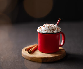 Red mug with hot chocolate or cocoa with whipped cream on a wooden stand with cinnamon sticks  on a dark background