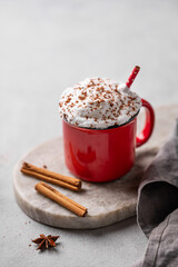 Red mug of hot cocoa or chocolate with whipped cream and cinnamon sticks on a gray background. The mug sits on an oval marble stand with a napkin. Free space for text.