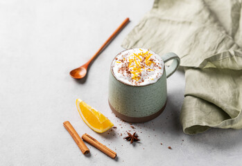 Mug of hot chocolate with whipped cream and orange zest on a light gray background with a cinnamon sticks, anise star, spoon and fresh orange slices.