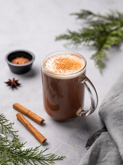 Obraz na płótnie Canvas Glass mug with hot chocolate and milk foam on a light background with cinnamon sticks, anise star and pine tree branches.