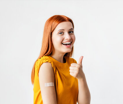 Portrait of a red-haired smiling young woman after getting a vaccine standing on a white background, showing her arm with bandage after receiving vaccination