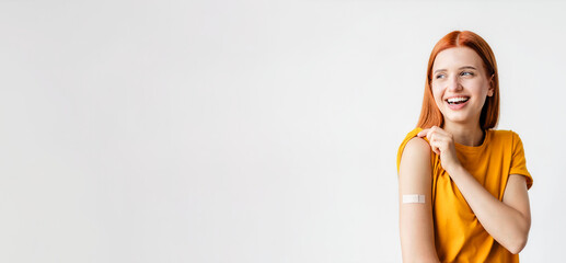 Portrait of a red-haired smiling young woman after getting a vaccine standing on a white background, showing her arm with bandage after receiving vaccination - obrazy, fototapety, plakaty