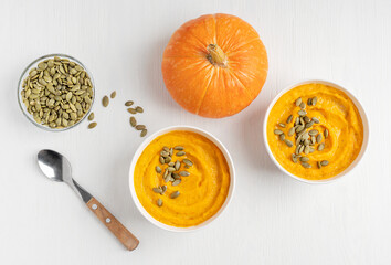 Top view of homemade creamy pumpkin soup made of ripe squash mashed or pureed with dairy cream and broth decorated with seeds served in bowls on white wooden background with spoon for healthy dinner