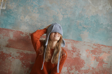 Laughing girl in sweater and beanie pulled down over eyes leaning against shabby wall