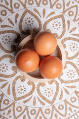 eggs, aerial photo of three eggs placed on a circular wooden bowl, the table has a tablecloth with mandalas in white and brown tone