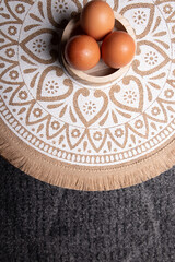 eggs, aerial photo of three eggs placed on a circular wooden bowl, the table has a tablecloth with mandalas in white and brown tone