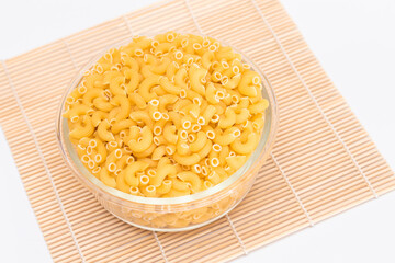 Uncooked Chifferi Rigati Pasta in Glass Jar on Bamboo Mat on White Background. Fat and Unhealthy Food. Classic Dry Macaroni. Italian Culture and Cuisine. Raw Pasta