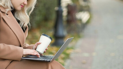 Online communication. Happy woman. Virtual life. Smiling lady sitting bench in park alley typing laptop holding cup of coffee.