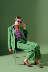  Fashionable beautiful confident woman wearing trendy color sunglasses, suit blazer, classic pants, purple office shirt, zebra print pointed toe shoes, posing on green background. Full-length portrait