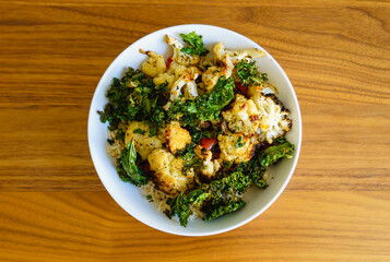 Vegetarian meal with oven roasted cauliflower, kale, and red bell peppers over brown rice in a white bowl on a wood table