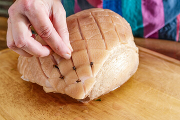 Hand of a woman larding the rind of a raw roast pork with cloves on a wooden kitchen board to cook...