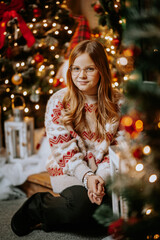 A young woman in a beige sweater sits in a Christmas decoration against the backdrop of a Christmas tree and lights. Girl on the porch