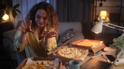 A young, curly-haired freelance woman dressed in a yellow shirt works at home at night. Her desk is lined with boxes of pizza and she enjoys the taste of it while working