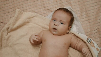 Small baby lies on bed, waves his hands, frowns.  Newborn baby lies on a bath baby towel, frowns. Undressed child of European appearance. Negative emotion. Blue eyes.  Newborn baby looks with interest