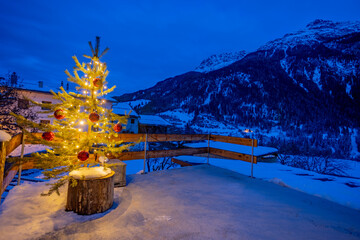 Real Christmas tree illuminated at dusk on the terrace of a house in a mountain village decorated with big red balls and lights all around. Big mountains on the background.