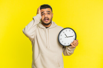 Indian young man with anxiety checking time on clock, running late to work, being in delay, deadline. Arabian Hindu guy looking at hour, minutes, worrying to be punctual isolated on yellow background