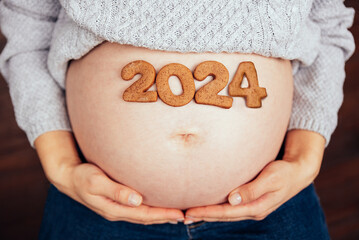 Cropped photo of pregnant woman holding number 2024 of ginger cookies on belly. people and expectation concept. Happy motherhood, healthy life. expecting a child. Baby born in 2024