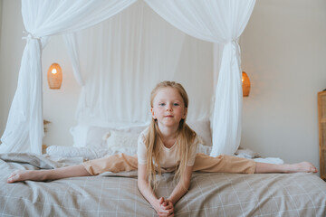 Little blonde girl in domestic clothes sitting on bed on splits at bedroom looks at camera,...