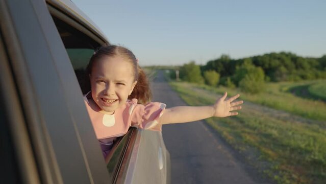 Child, stretching his hand out of car window, laughs, kid emotions. Girl child looks out of car window. Happy family travels by car. Little girl rejoices in family train by car. Family vacation