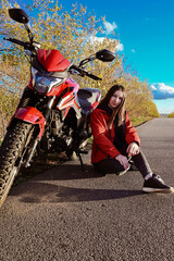 Obraz premium young attractive biker girl in red sitting next to her red motorcycle in the middle of the road. biker with a motorcycle outside the city.
