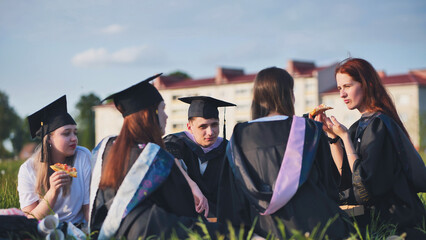 Graduates in black suits eating pizza in a city meadow.