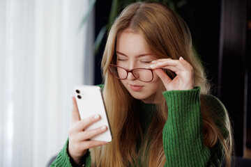 girl squinting with poor eyesight wearing glasses looking at a mobile phone at the table, concept...