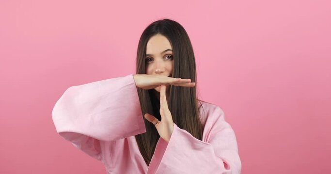 Portrait of displeased young caucasian woman showing time out hand gesture, looking imploringly, worried about deadline, wearing pink dress, posing isolated on pink color background wall in studio.