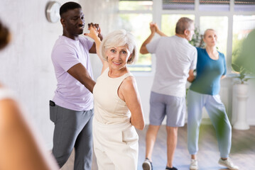 Fototapeta premium Happy smiling elderly woman enjoying impassioned merengue with african american partner in latin dance class. Social dancing concep