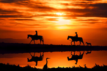 two horse riders in front of a beautiful sunset with a dog trailing behind - 684367623