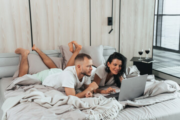 Obraz na płótnie Canvas Young caucasian couple laying on bed with laptop celebrating anniversary choosing trip via internet. Travel, leisure activities, honeymoon. Happy newlyweds at hotel apartments relaxing.