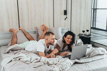 Fototapeta premium Young caucasian couple laying on bed with laptop celebrating anniversary choosing trip via internet. Travel, leisure activities, honeymoon. Happy newlyweds at hotel apartments relaxing.