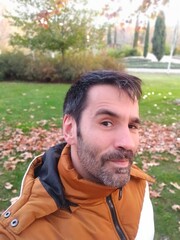 Selfie portrait of Caucasian man looking at camera with short dark hair in brown and white coat suspicious with raised eyebrow and looking at camera in a park during autumn winter - 684373412